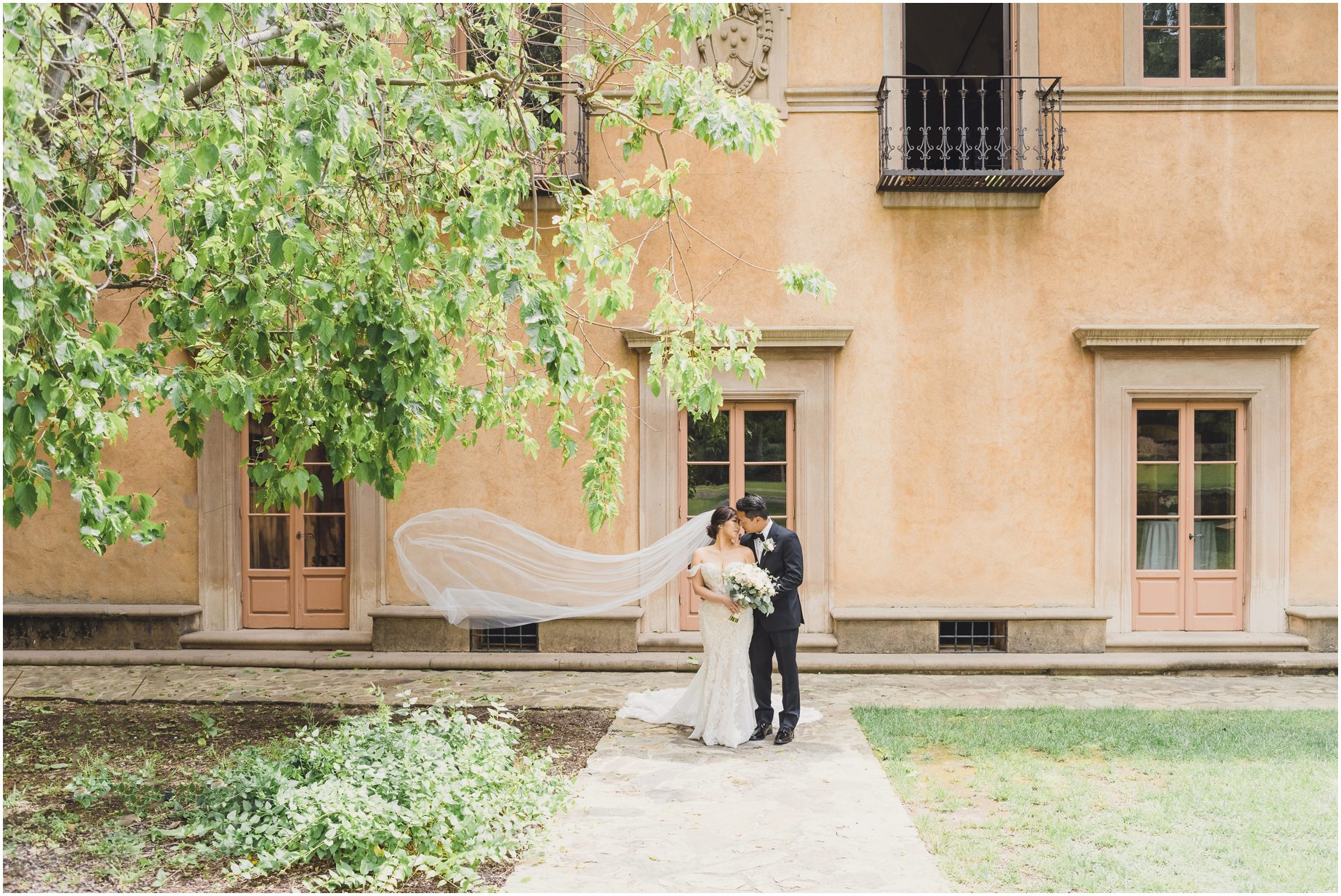 A bride and groom stand for a dreamy portrait in front of a building at Villa del sol d'Oro as the bride's veil floats in the breeze.