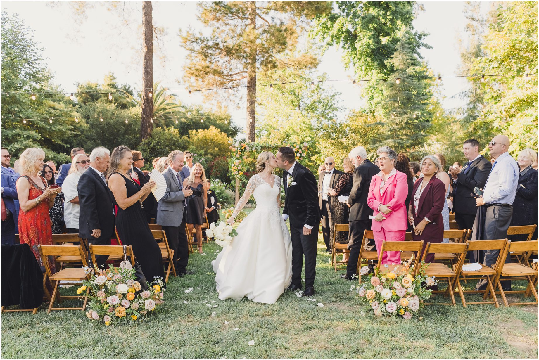As the bride and groom kiss, walking out of their wedding, their guests look on with joy. Colorful flowers flank the aisle, and trees surround the background of the ceremony space. The lodge at Malibou lake is a dreamy location for a wedding