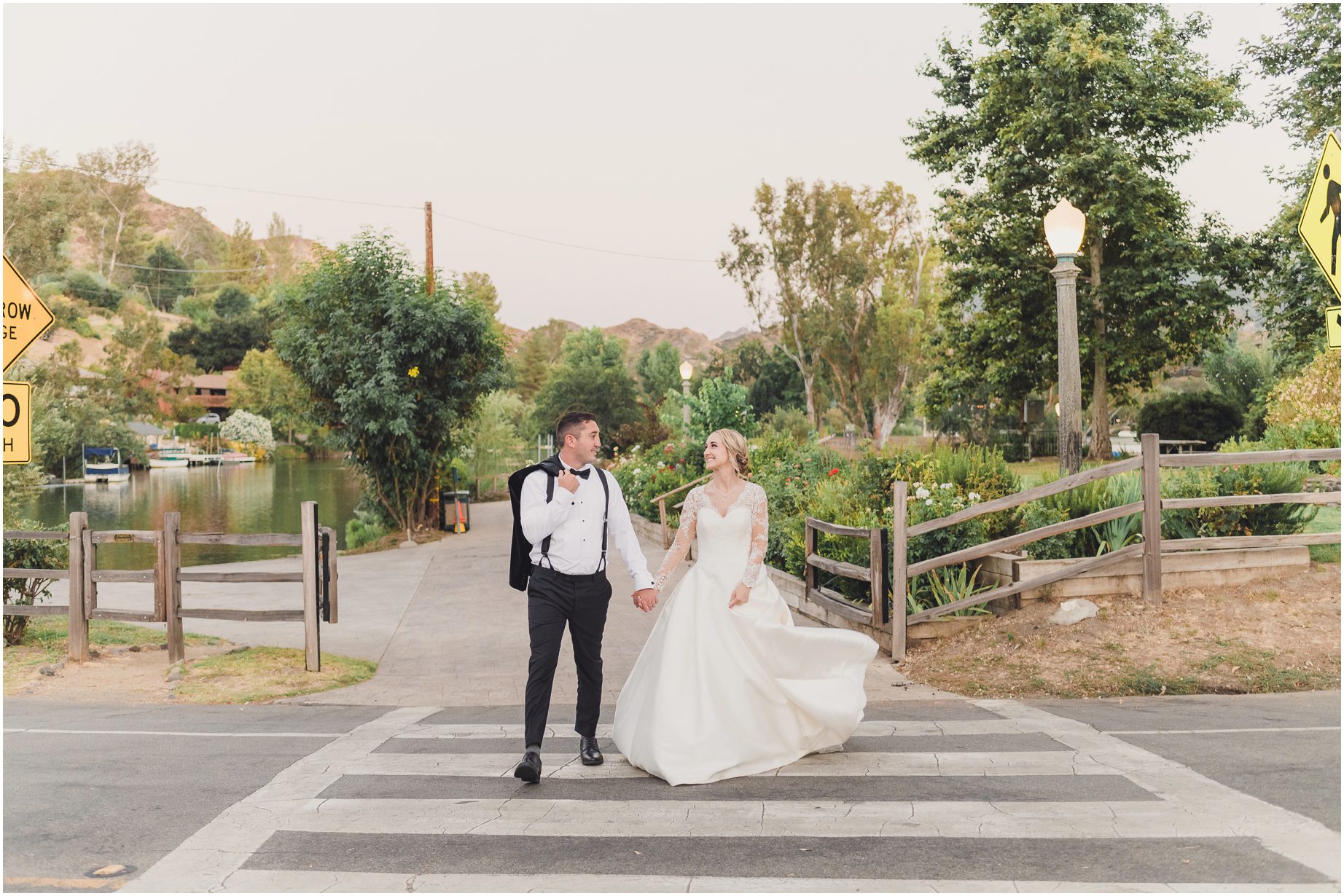 The bride and groom cross the street at the Lodge at Malibou Lake near the lake. They are smiling at each other and the streetlights have just come on because the sun is setting