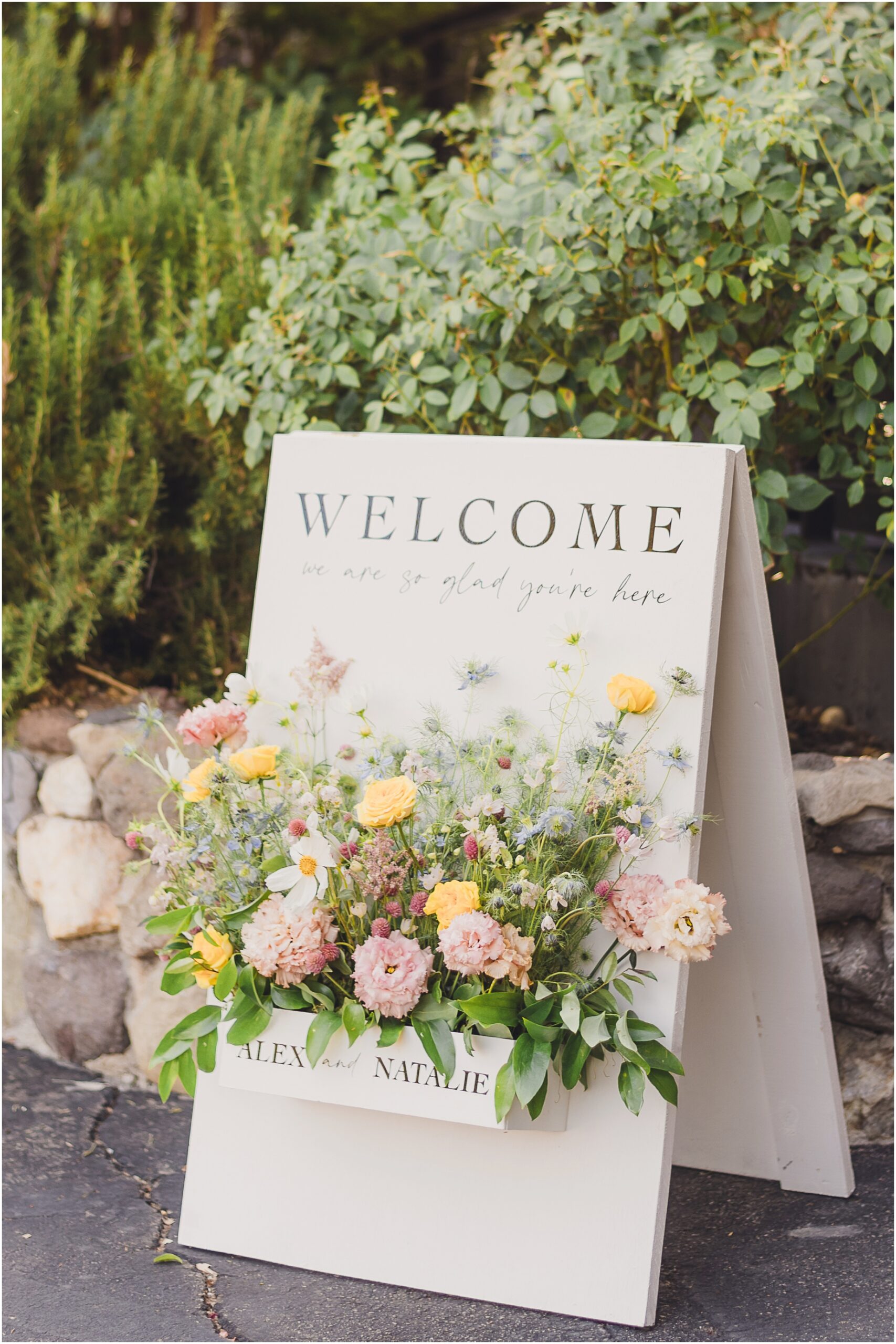 A welcome sign greets people entering Alex and Natalie's wedding at the lodge at Malibou Lake in Southern California. The sign is adorned with colorful and tasteful flower arrangements