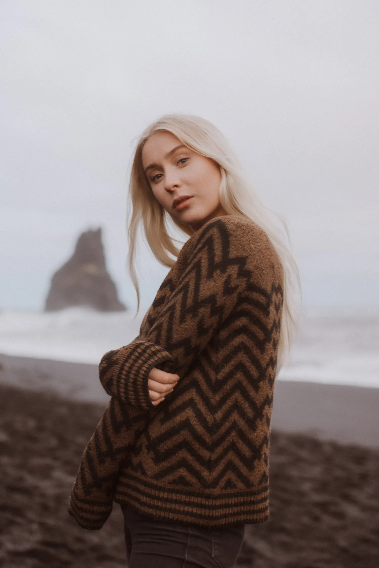 A woman with platinum blonde hair wearing a chevron-patterned sweater at a black sand beach, gazing over her shoulder with a serene expression, with a rugged sea stack blurred in the background.