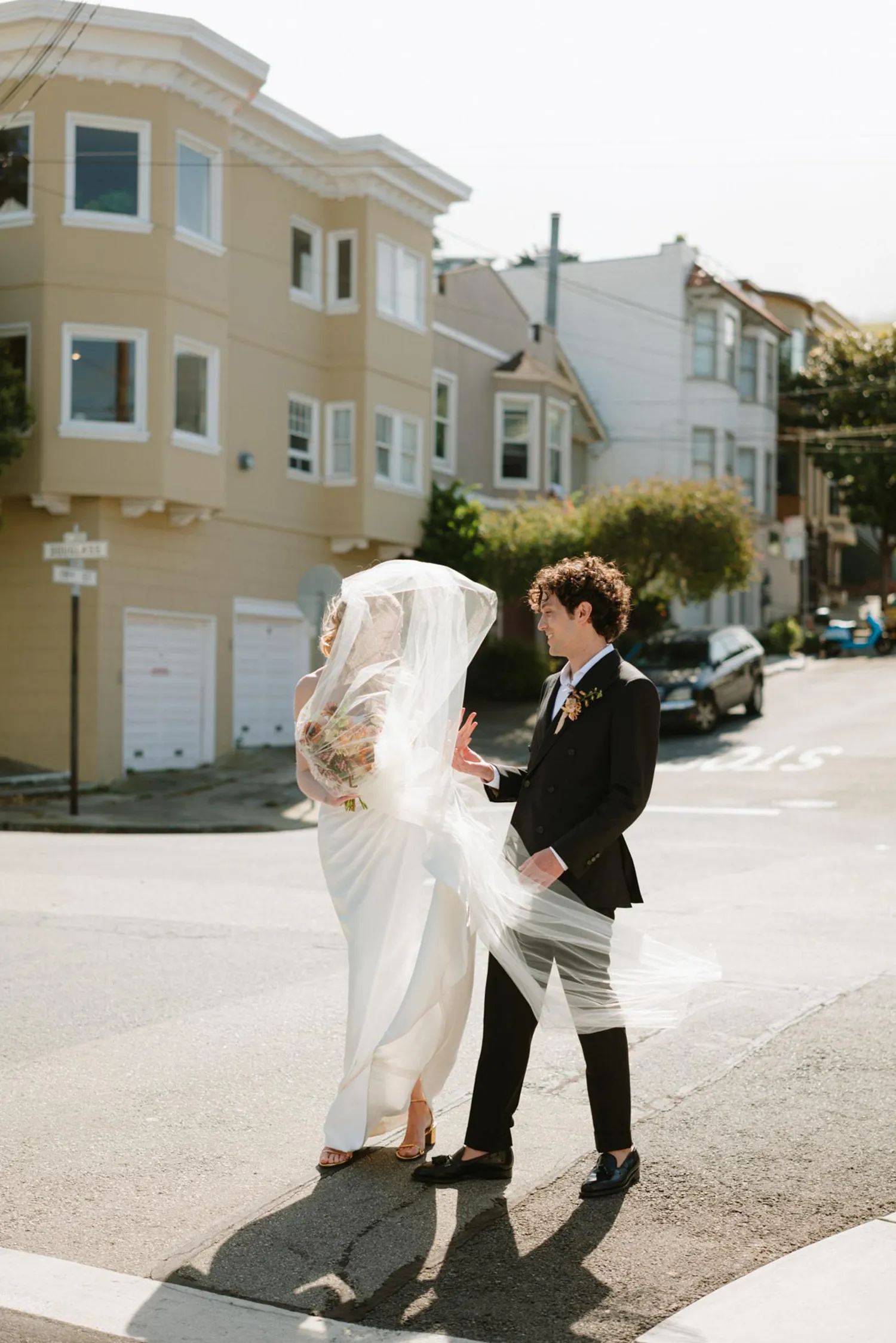 Top Wedding Photographers of 2024, featuring Lukas Korynta's work: A bride in a flowing dress with a delicate veil walks with her groom in a sharp suit across a city street, with classic San Francisco houses framing this candid, sunlit moment.