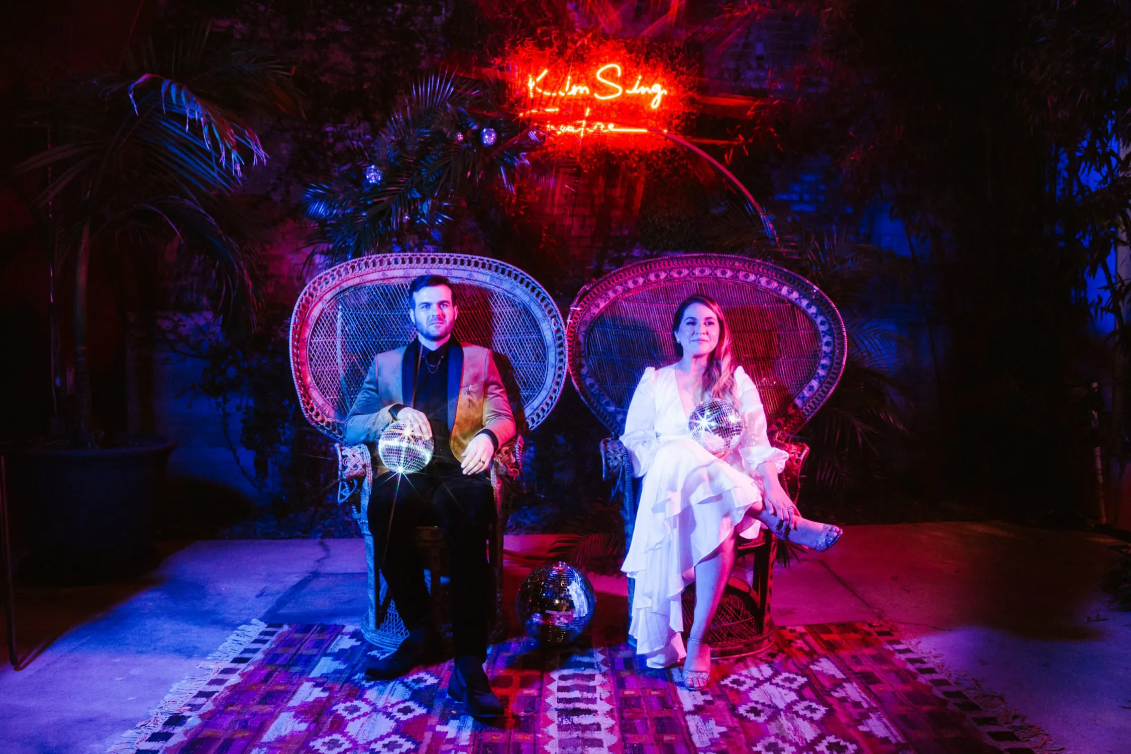 A man and woman seated on peacock chairs under a neon 'Kim Sing Theatre' sign, bathed in vibrant blue and red lighting, creating a dynamic and stylish atmosphere at a trendy event venue.