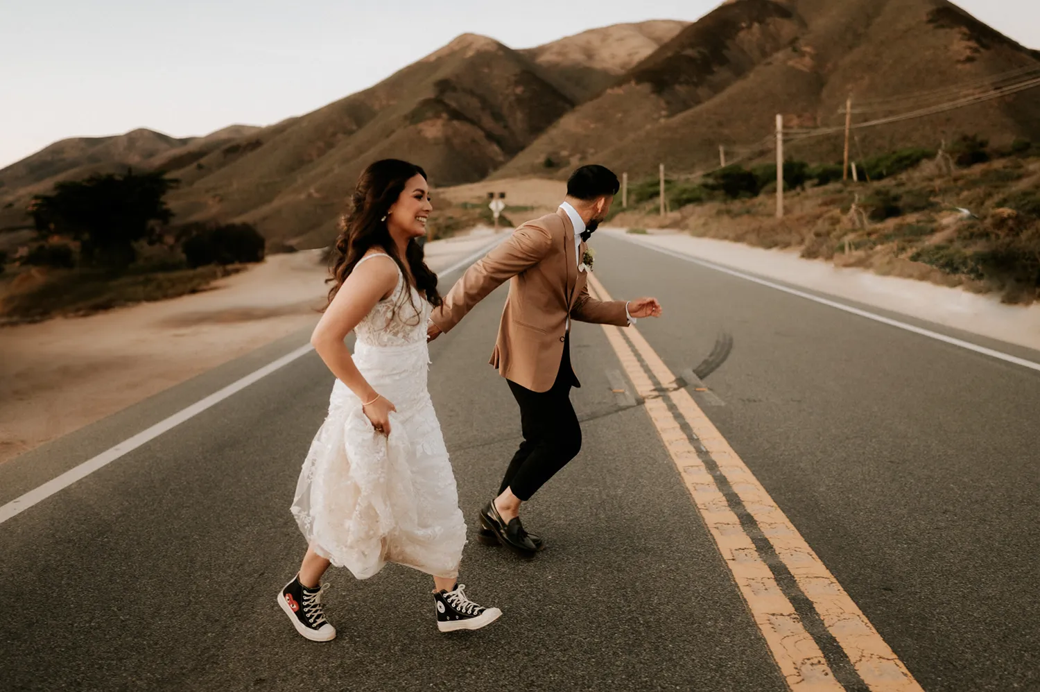 Captured by Heather K Purdy, a name among the Top Wedding Photographers of 2024, a bride and groom in casual footwear dash across a deserted road, with the rolling hills of California as their backdrop, reflecting a spontaneous and adventurous spirit.