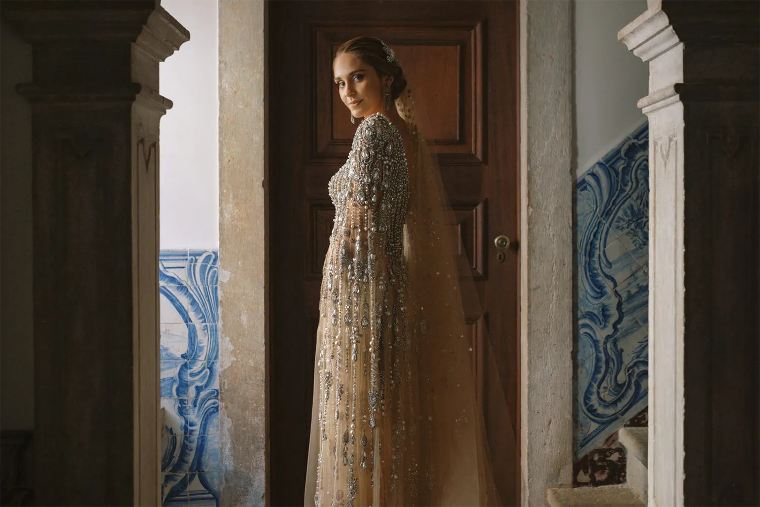 Top Wedding Photographers of 2024 capture by Nirav Patel features a bride in a bejeweled golden gown standing by a wooden door, with traditional Portuguese azulejos tiles in the background, creating an air of timeless elegance.