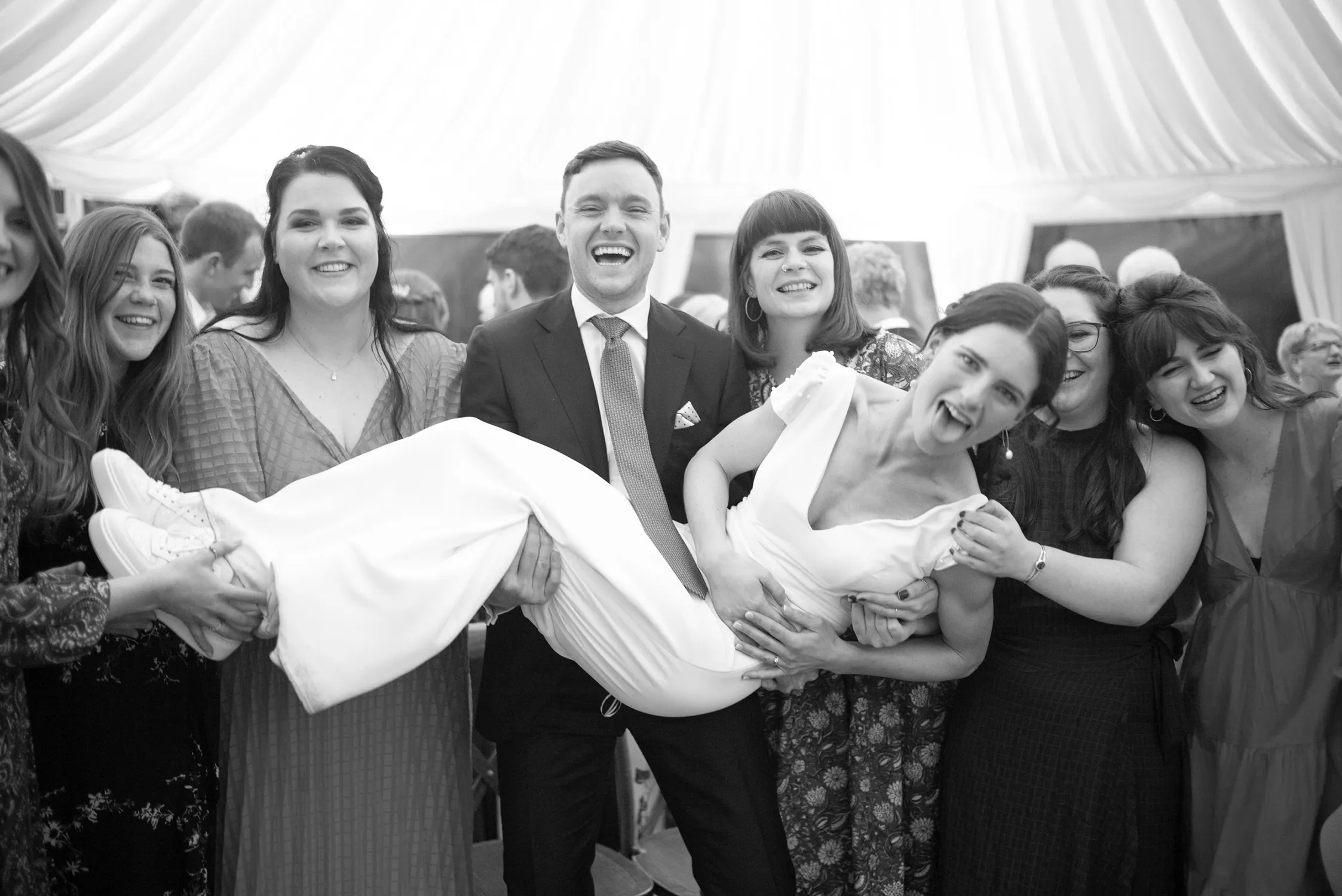 A jubilant wedding scene photographed by John Dolan, distinguished in the Top Wedding Photographers of 2024, captures a groom carrying his bride amidst a group of laughing friends, all sharing in the couple’s joy on their big day.