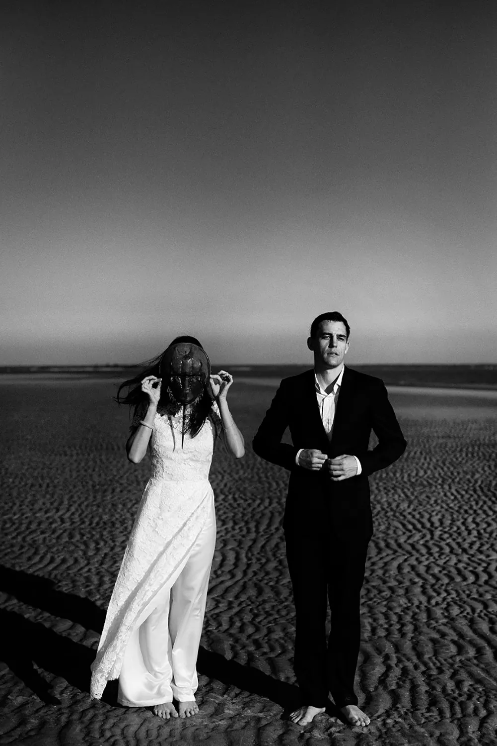 A striking monochrome portrait by Adonye Jaja, celebrated among the Top Wedding Photographers of 2024, features a bride and groom standing apart on a textured sand dune, the bride adjusting her veil and the groom looking pensively ahead.