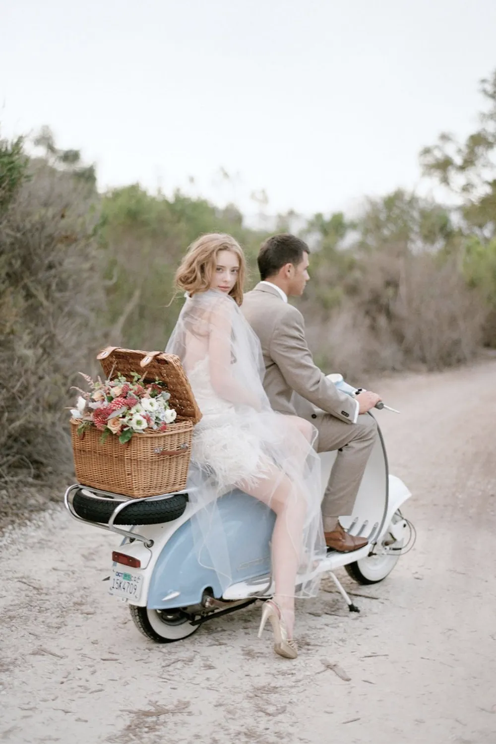 A whimsical escape captured by Elizabeth Messina, an elite name in the Top Wedding Photographers of 2024, featuring a bride and groom on a classic scooter with a wicker basket full of flowers, set against a soft, natural backdrop, exuding a vintage romance.