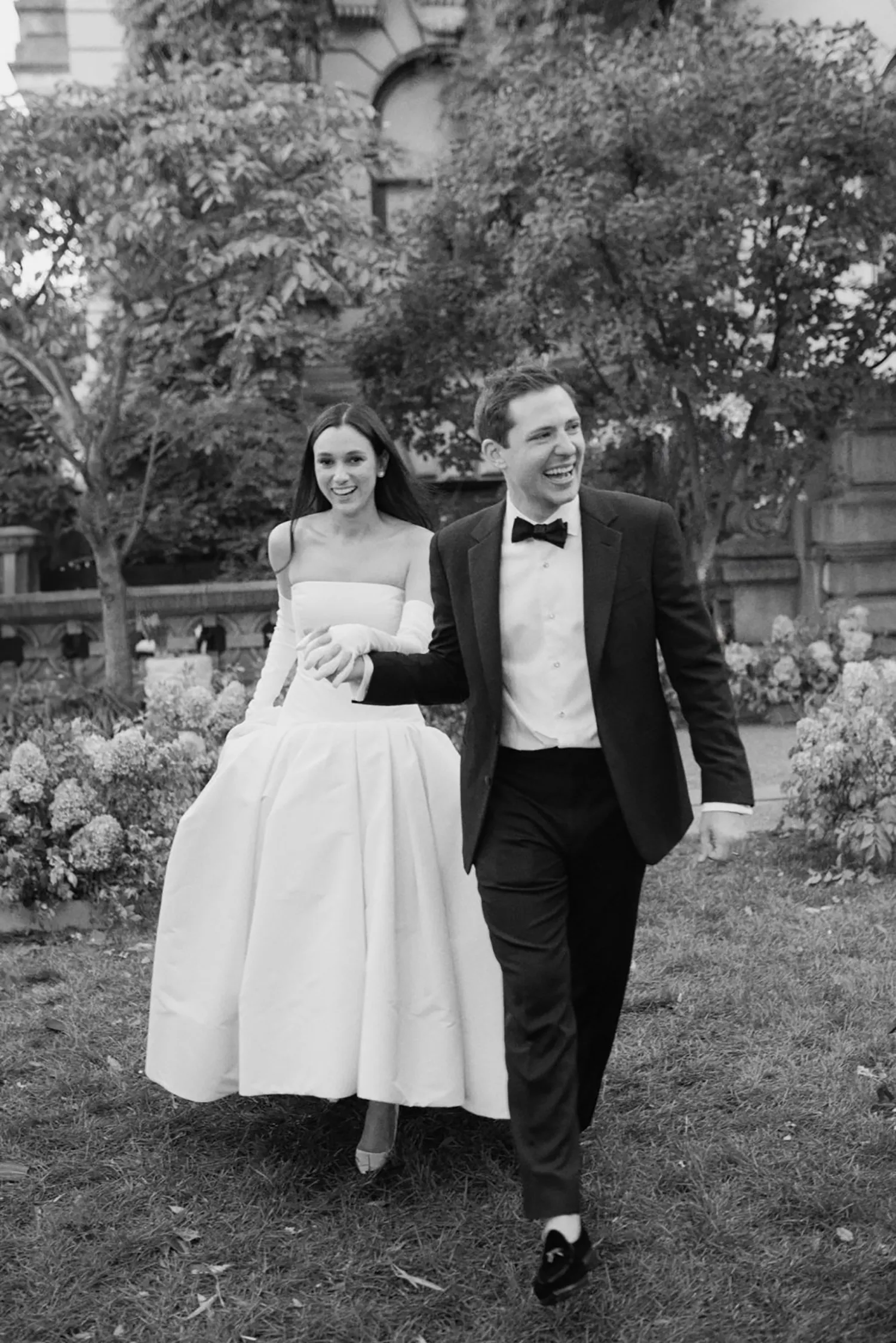 A candid moment of joy by Samm Blake, recognized in the Top Wedding Photographers of 2024, depicts a chic bride in an elegant mid-length dress and a groom in a classic tuxedo, both laughing and walking hand in hand through a lush garden.