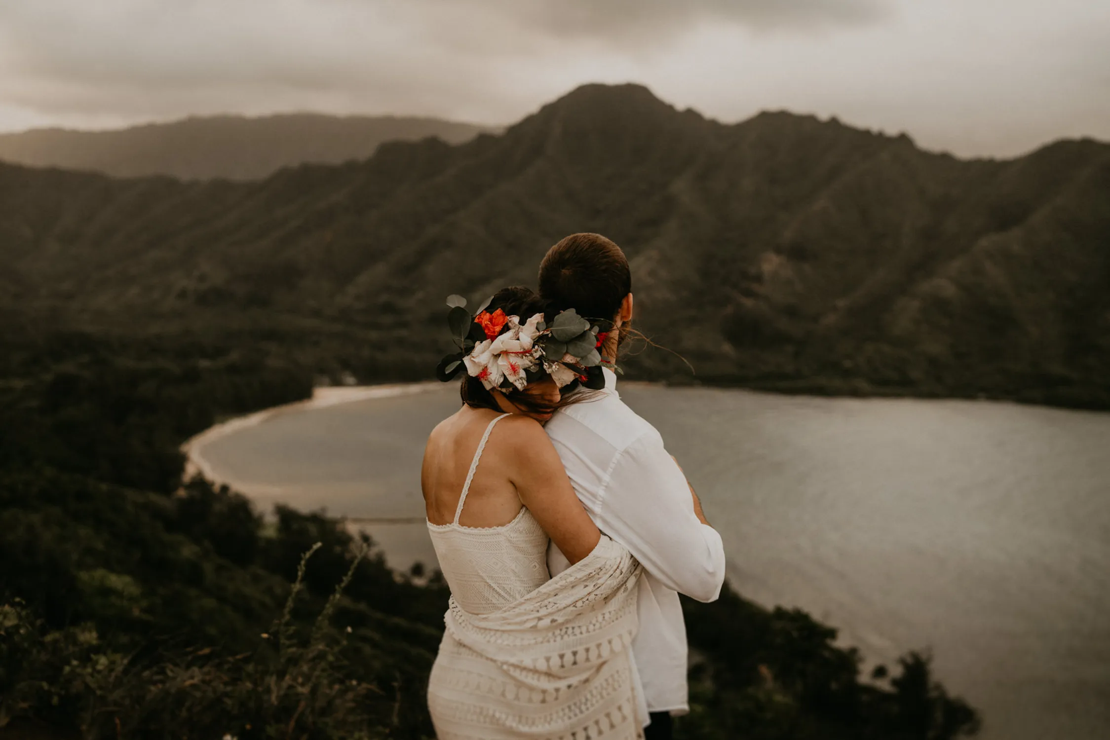 A serene scene captured by Henry Tieu, among the Top Wedding Photographers of 2024, depicts a bride with a vibrant floral crown embracing her partner, with the sweeping panorama of a mountainous landscape and tranquil bay behind them, evoking a sense of intimate connection in a grand natural setting.